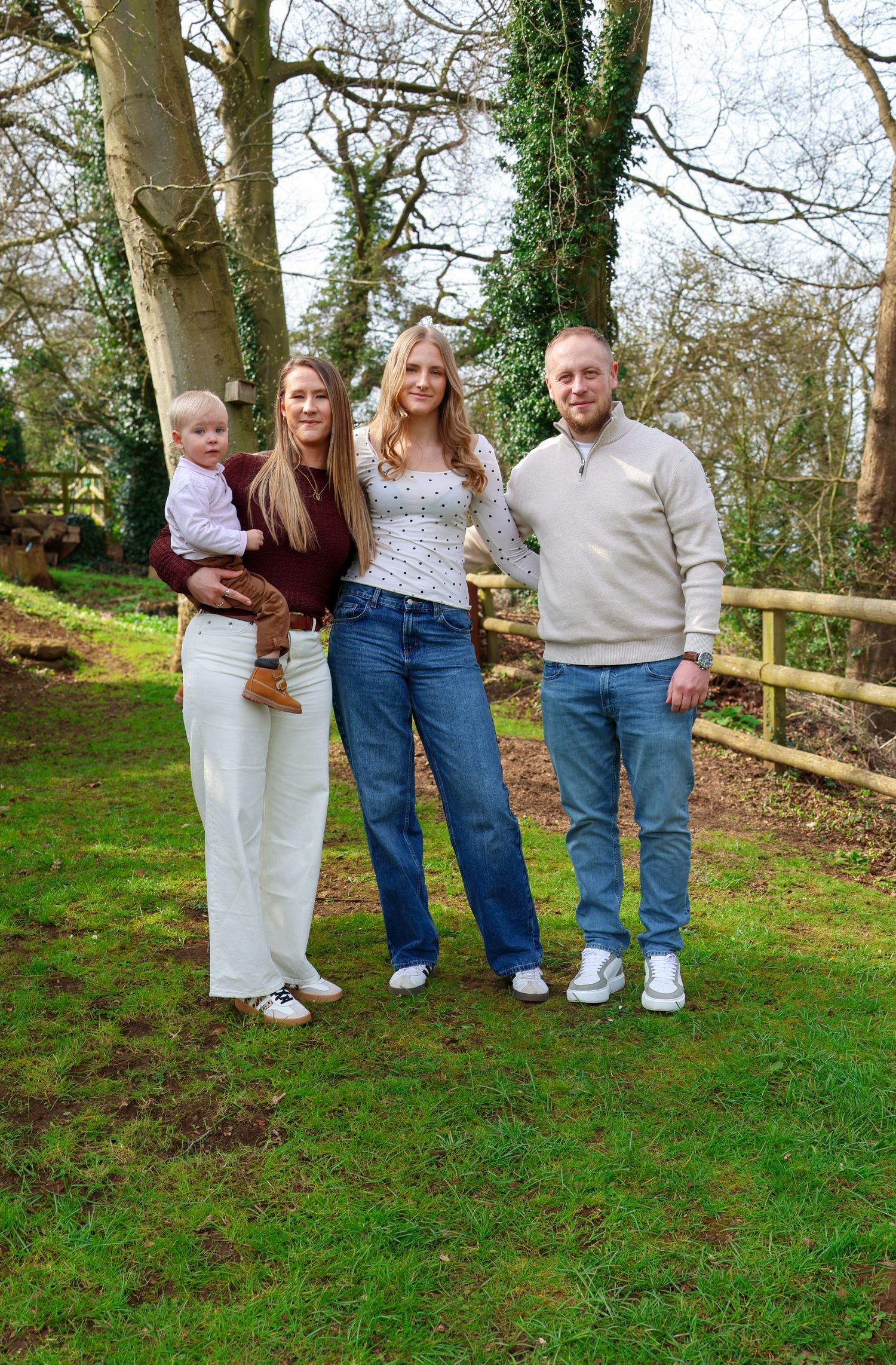 Family of four posing outdoors in a woodland garden setting, with parents holding a toddler on a green lawn.