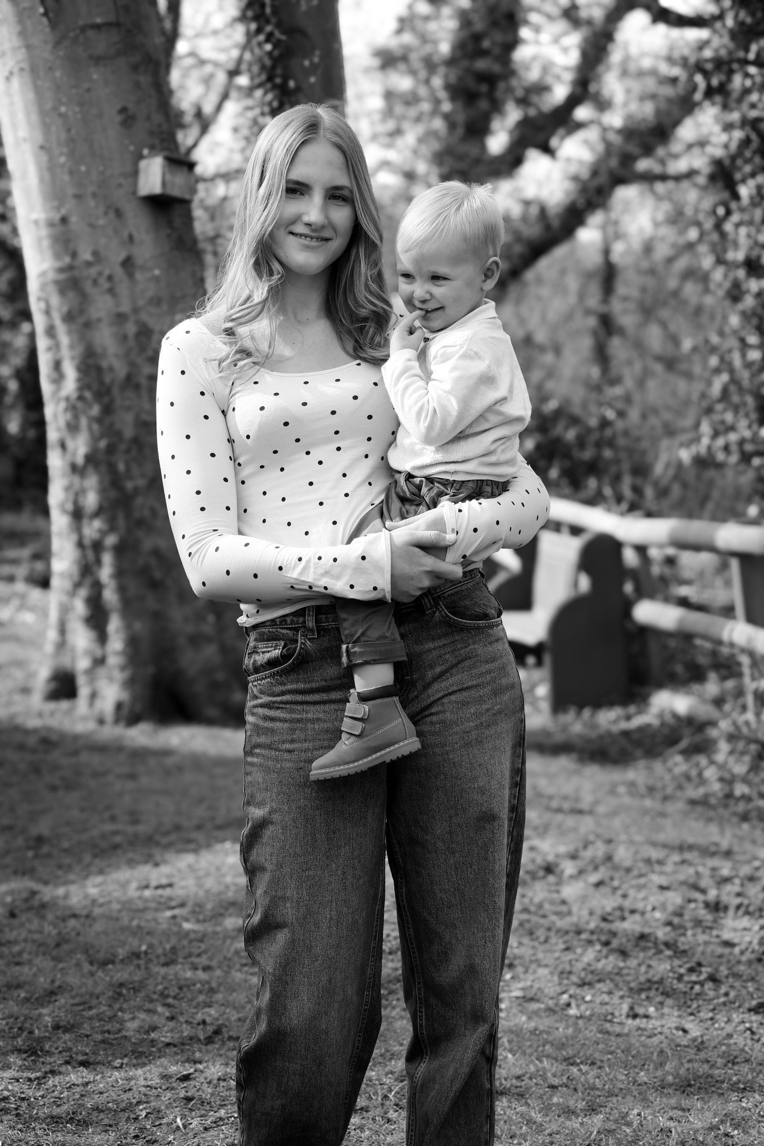 Black and white photo of a smiling blonde woman holding a toddler outdoors near trees.