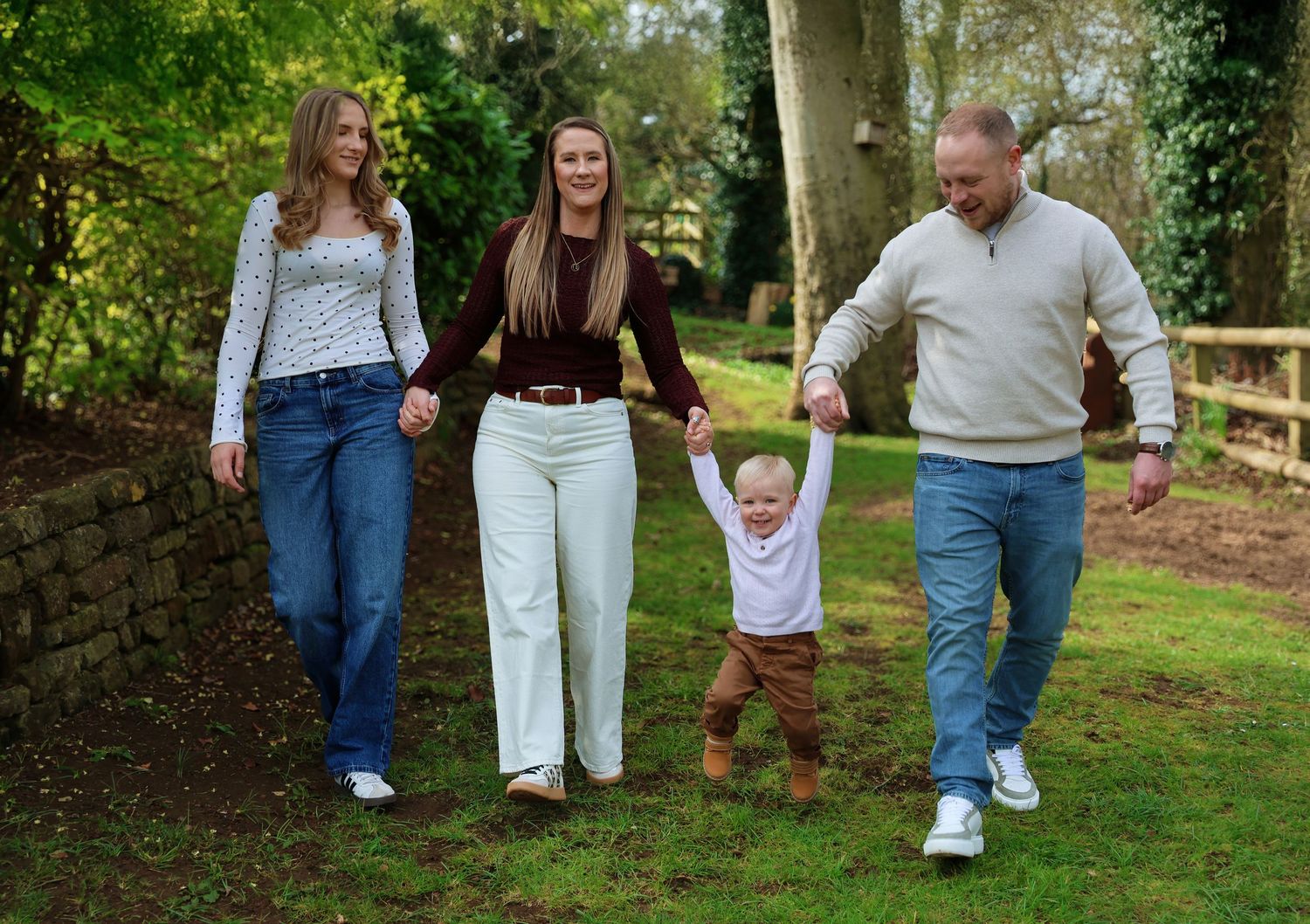Family of four walking in a park, parents swinging toddler by hands, surrounded by lush greenery.