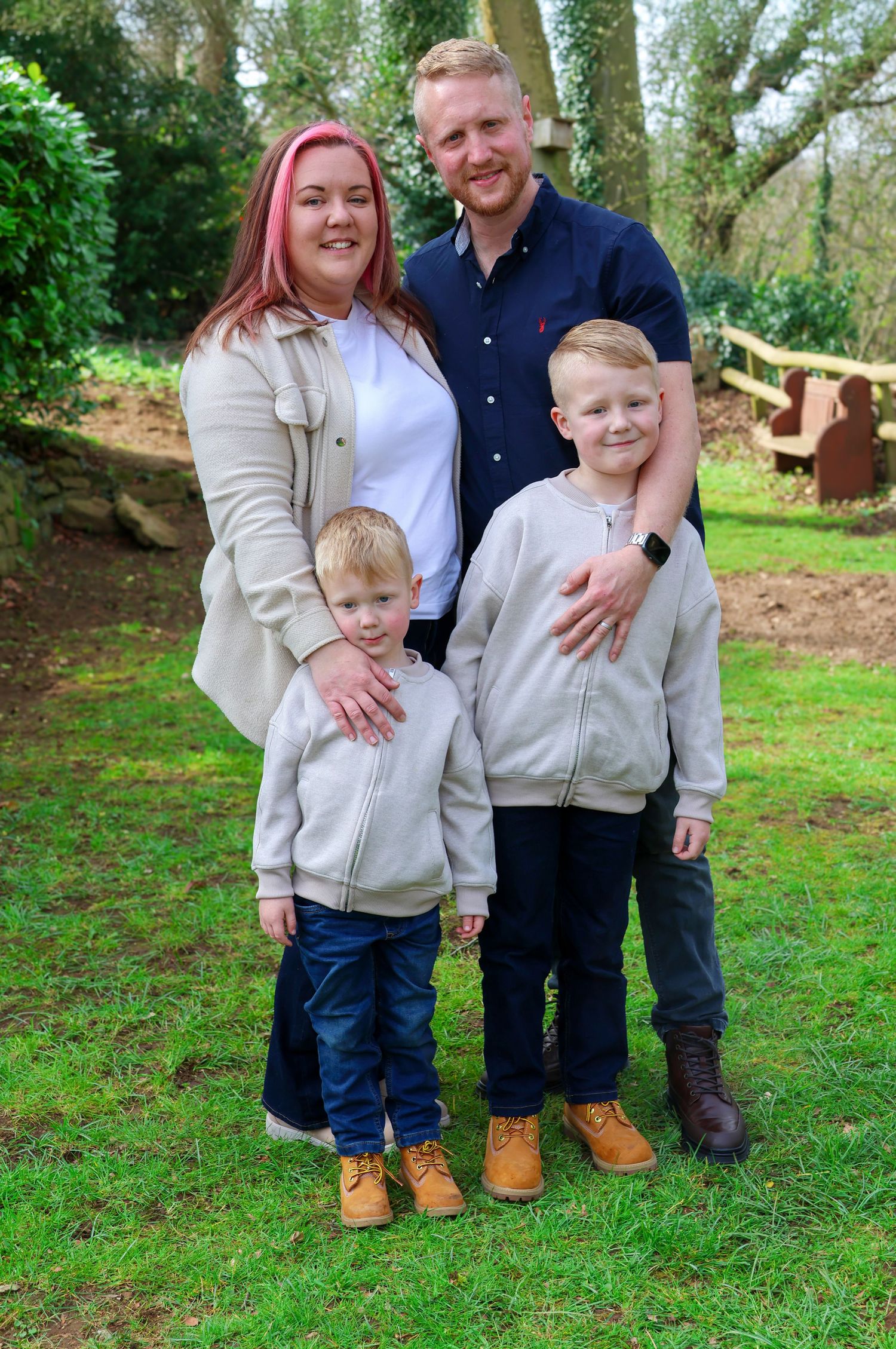 Family of four posing outdoors on grass, two adults and two young boys in matching grey hoodies and tan boots.