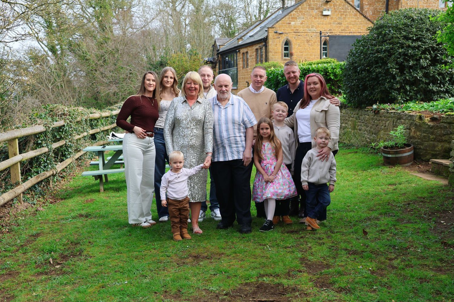 Multi-generational family group posing together outdoors in a garden setting near a wooden fence and stone building.