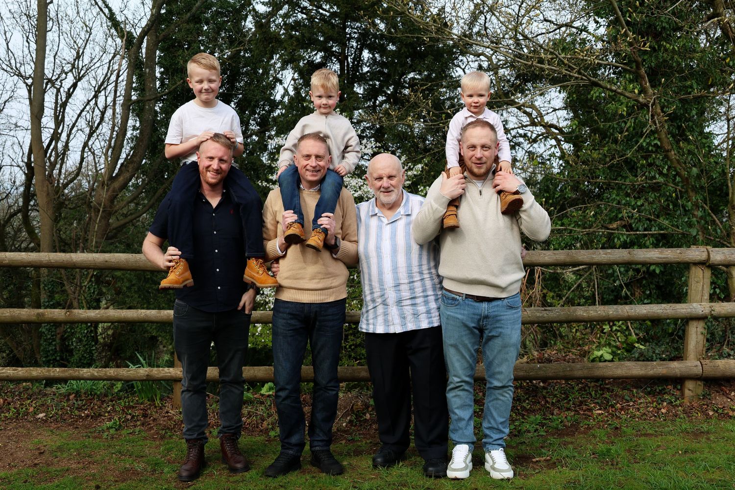 Three generations of men pose outdoors, with young children on shoulders, holding wooden crafts near a rustic fence.