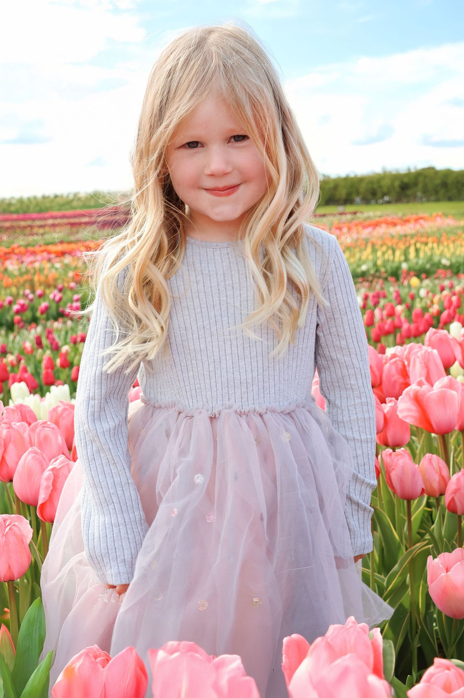 Young blonde girl in pink tulle dress smiling in a vibrant tulip field under a blue sky.