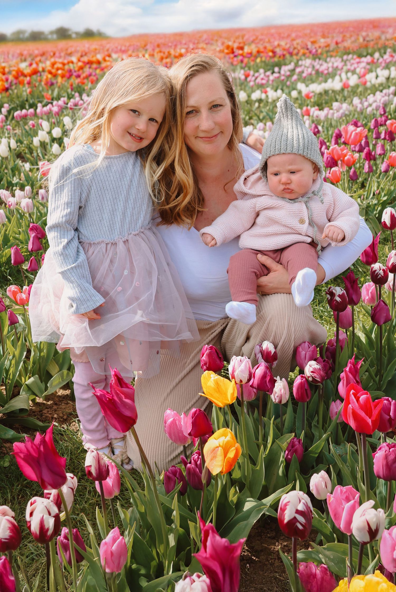 A woman and two young girls pose in a vibrant tulip field filled with pink, yellow, and orange blooms on a sunny day.