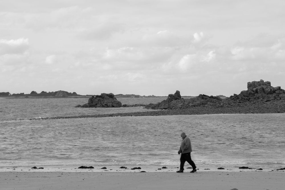 A silhouetted figure walks along a rocky beach shoreline in black and white photography.