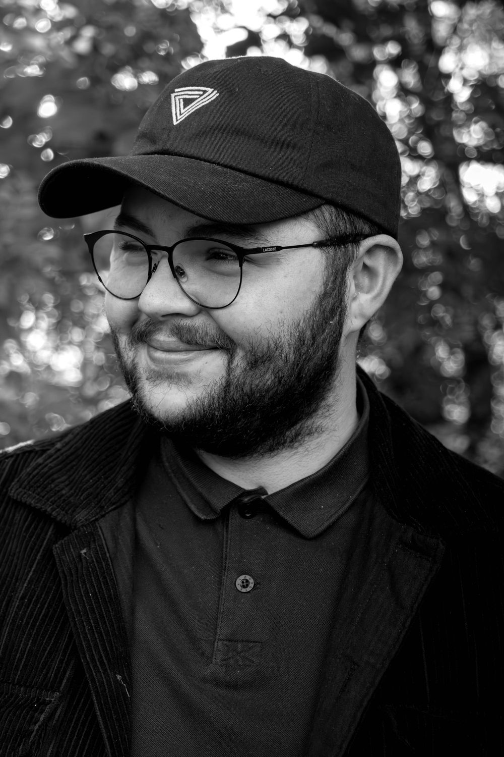 A black and white portrait of someone wearing glasses, a baseball cap with a geometric logo, and a black shirt outdoors.