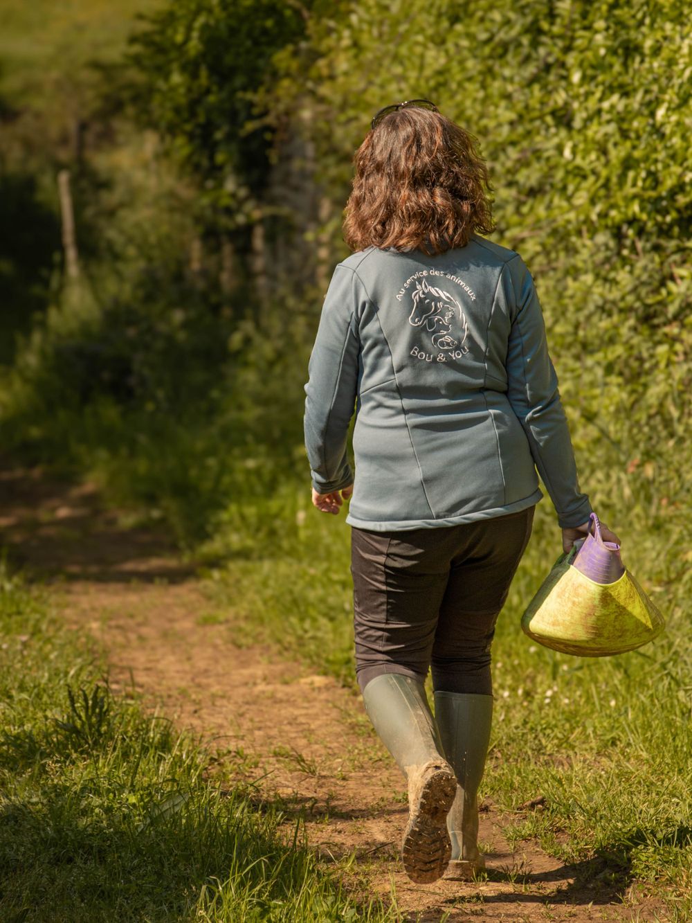 Person wearing rubber boots walks along a dirt path carrying a yellow bag in a sunny outdoor setting.