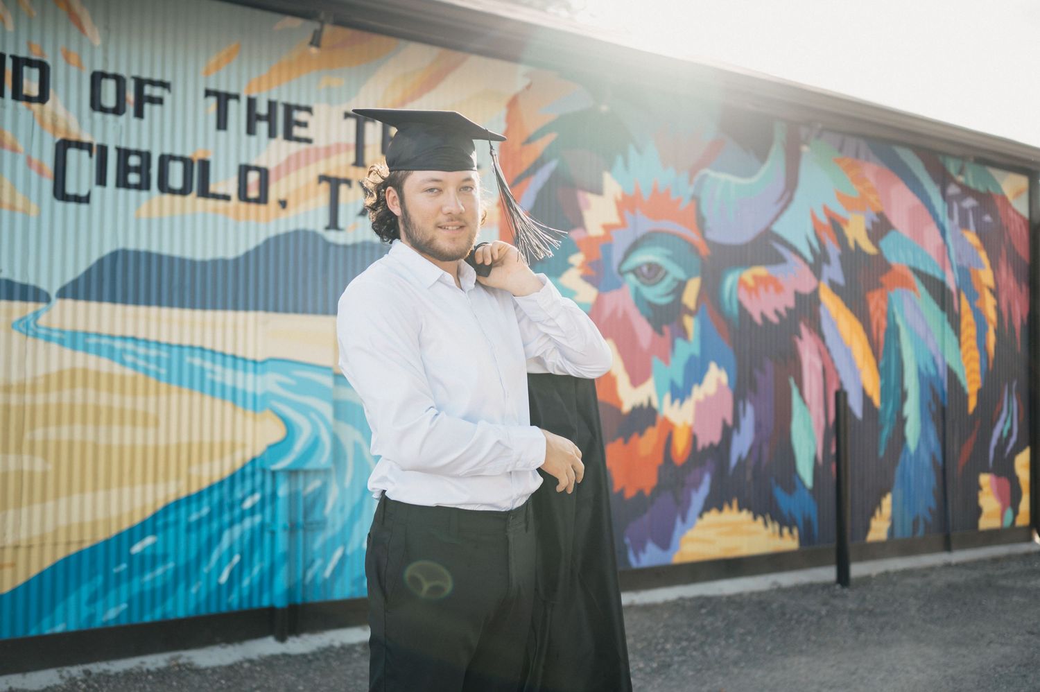 Graduate in cap and gown poses in front of vibrant street art mural with waves and colorful designs.