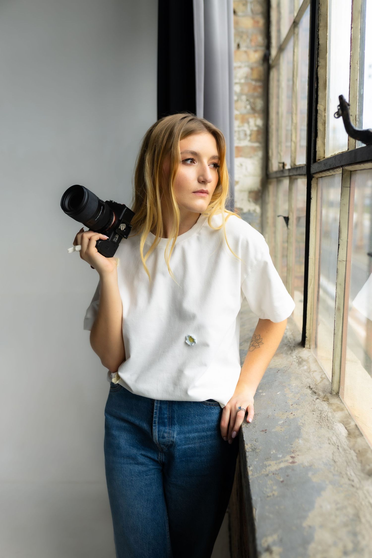 Photographer in casual white t-shirt and jeans holding camera by industrial window.