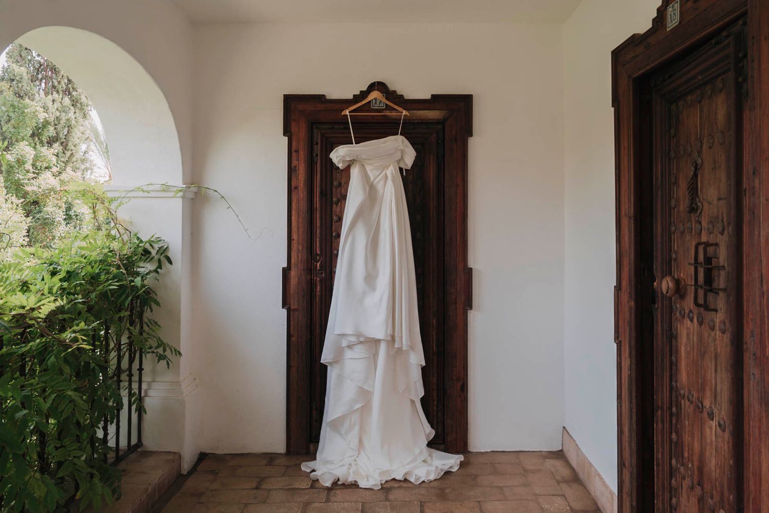 Elegant white wedding dress hanging on wooden wardrobe in rustic Mediterranean hallway with arched doorways.