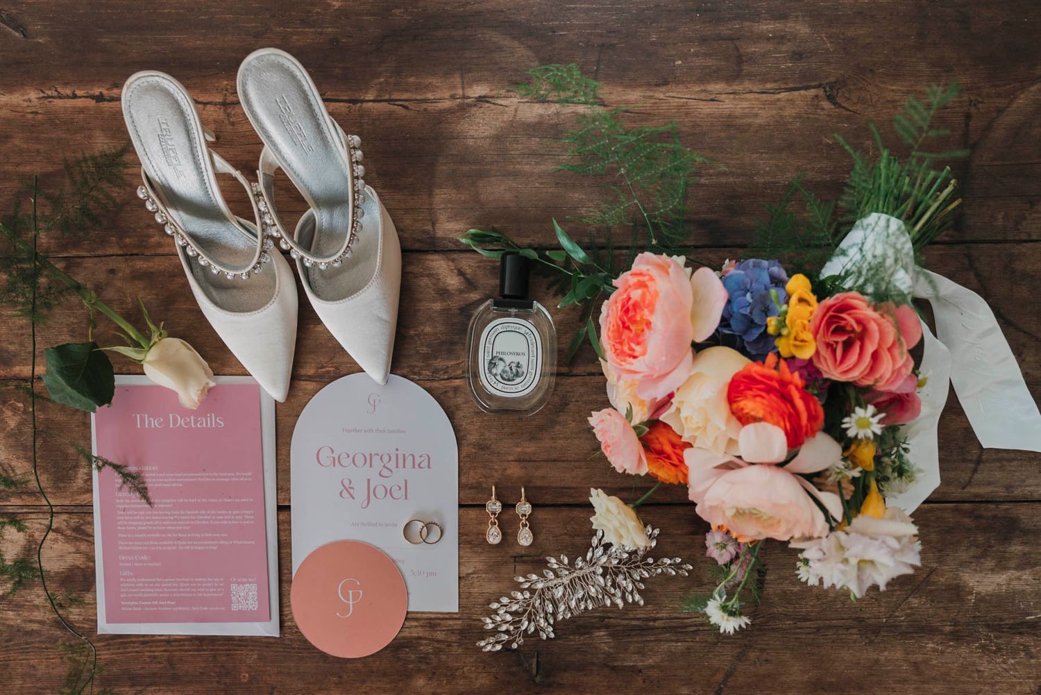 Wedding details flatlay with white shoes, invitation cards and colorful floral bouquet on wooden background.