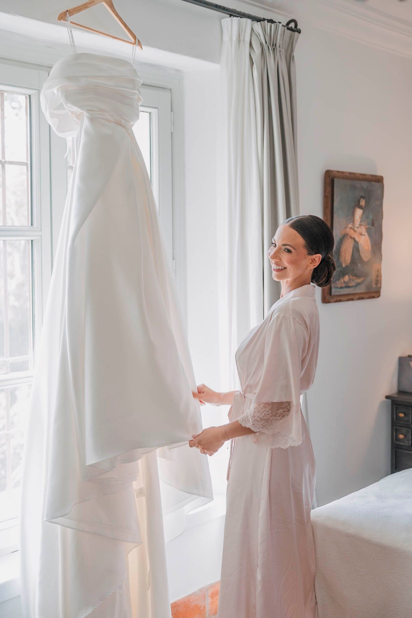 Elegant white wedding dress hanging by window while person in robe admires it.