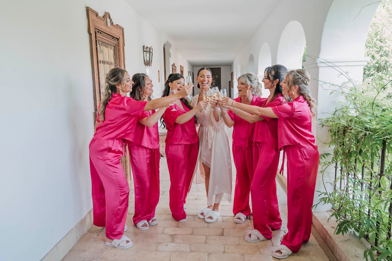 Bridesmaids in matching pink silk robes walk down a sunny corridor with the bride in white on her wedding day.