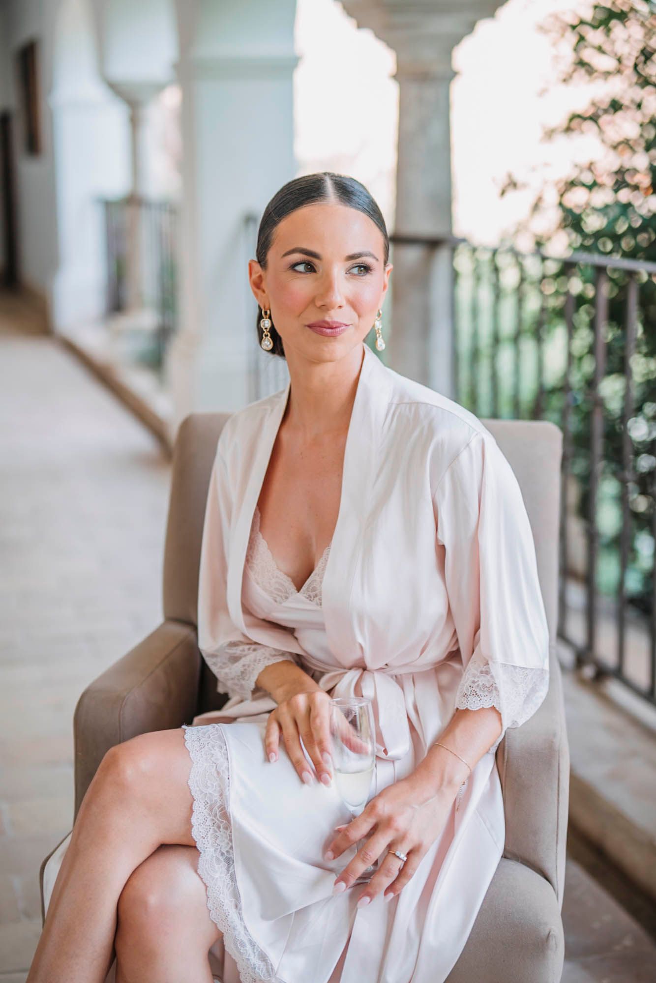 Person in white silk robe sits elegantly in chair on balcony during wedding preparation photoshoot.