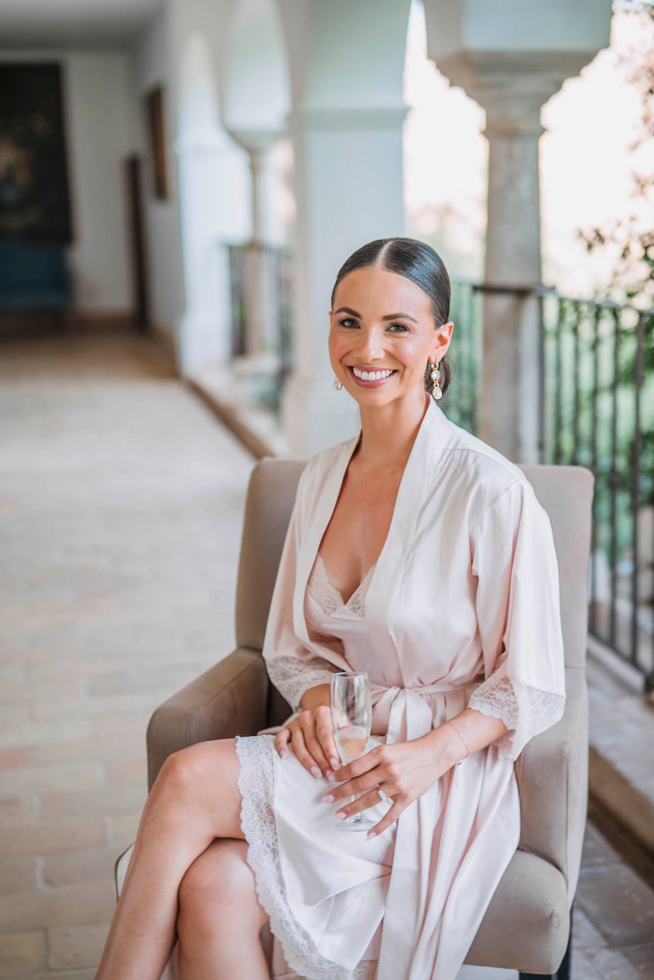 Person in white silk robe smiles while sitting in armchair during pre-wedding preparations.