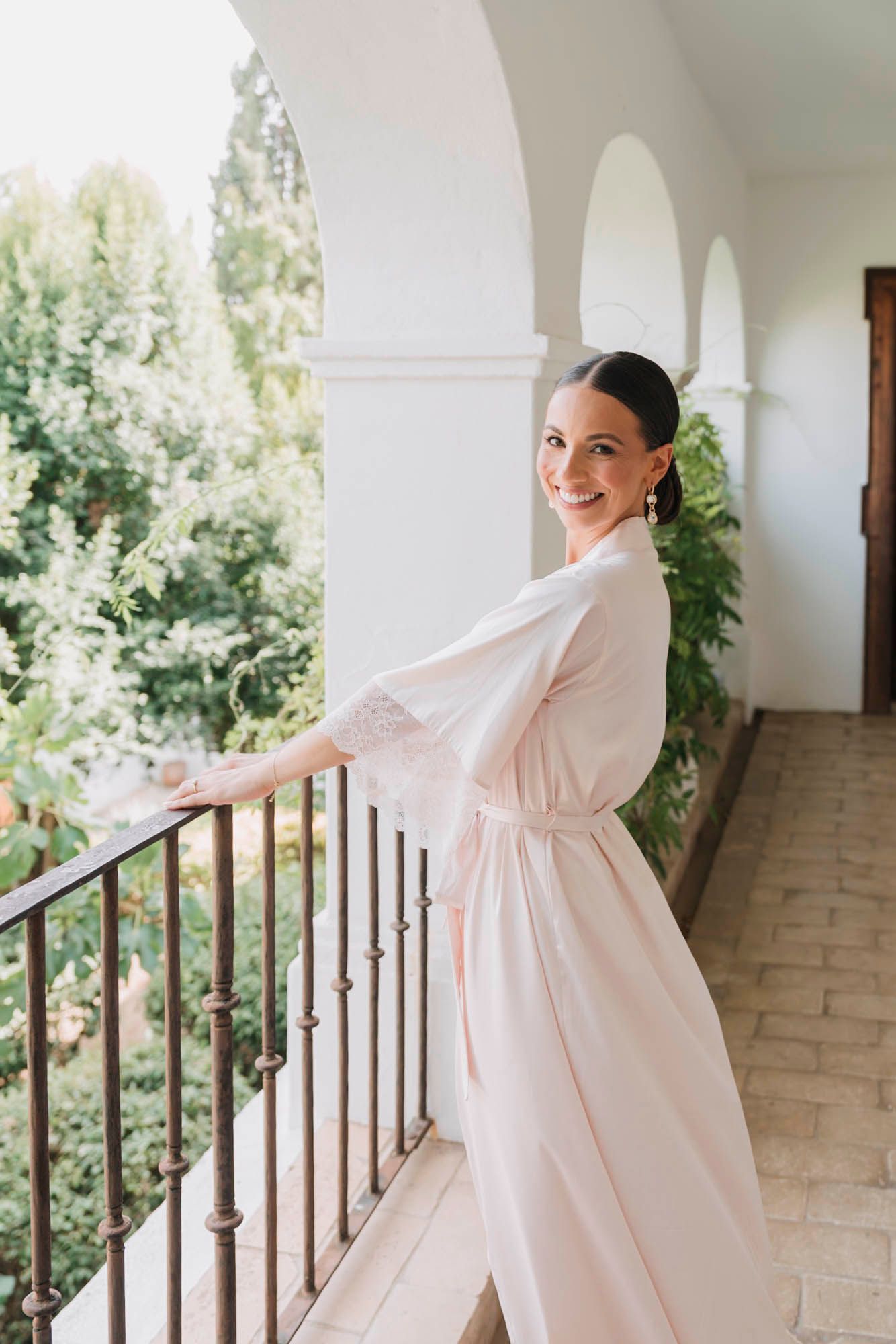 Person in white silk robe poses gracefully by balcony railing during wedding preparations.