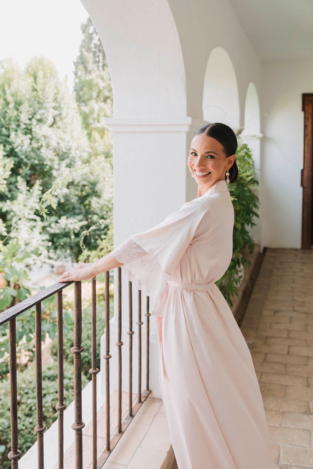 Person in white silk robe poses gracefully by balcony railing during wedding preparations.