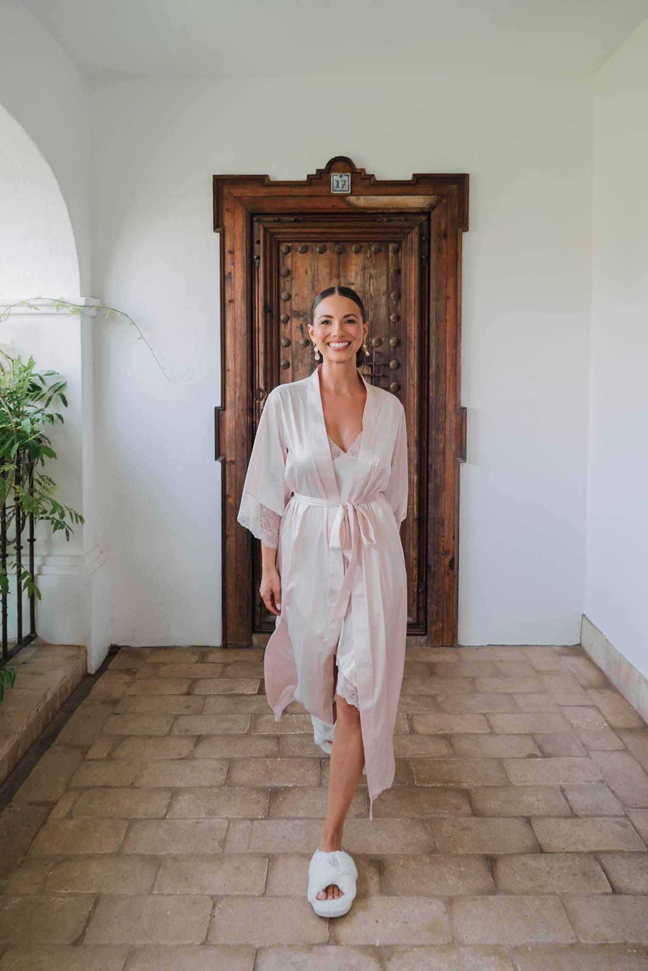 Person in white silk robe walks through arched hallway with wooden door backdrop.