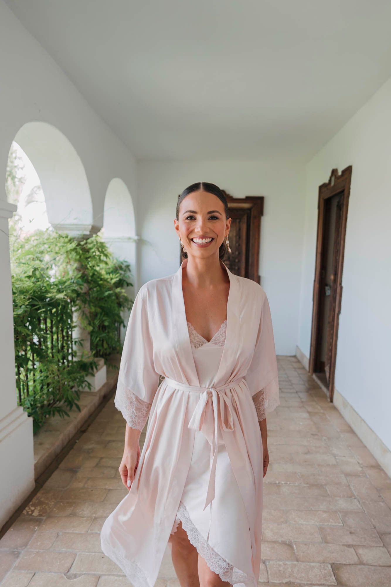 Person in flowing white robe smiles while standing in bright hallway with arched windows.