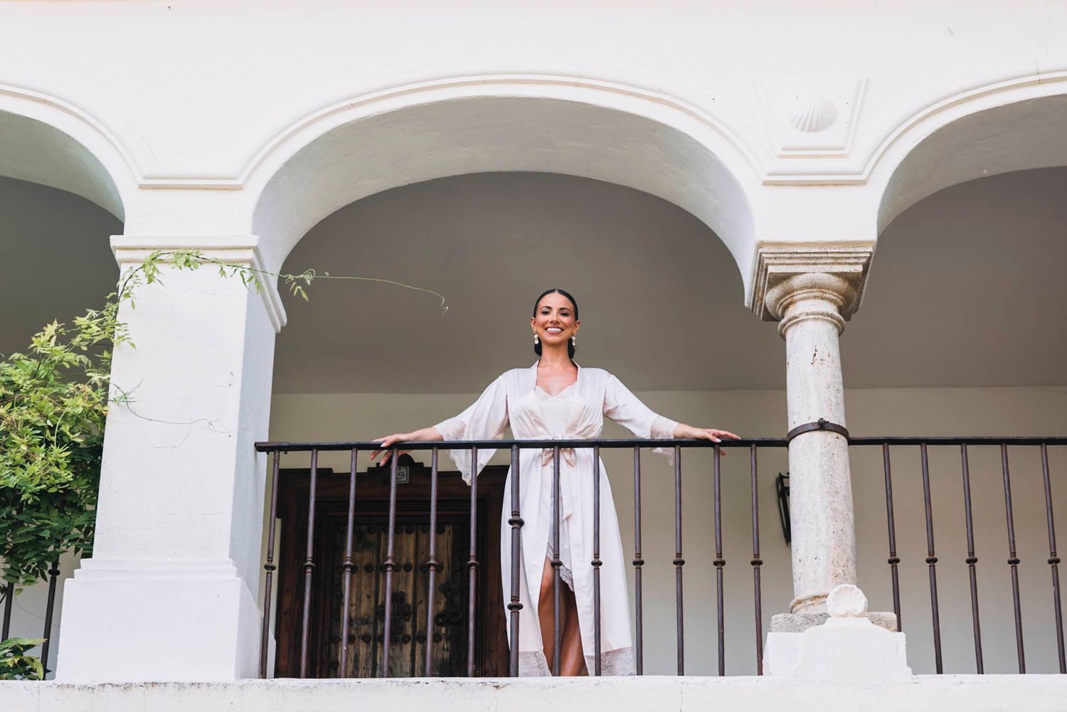 Person in white robe poses on balcony with classical architecture and arched columns.