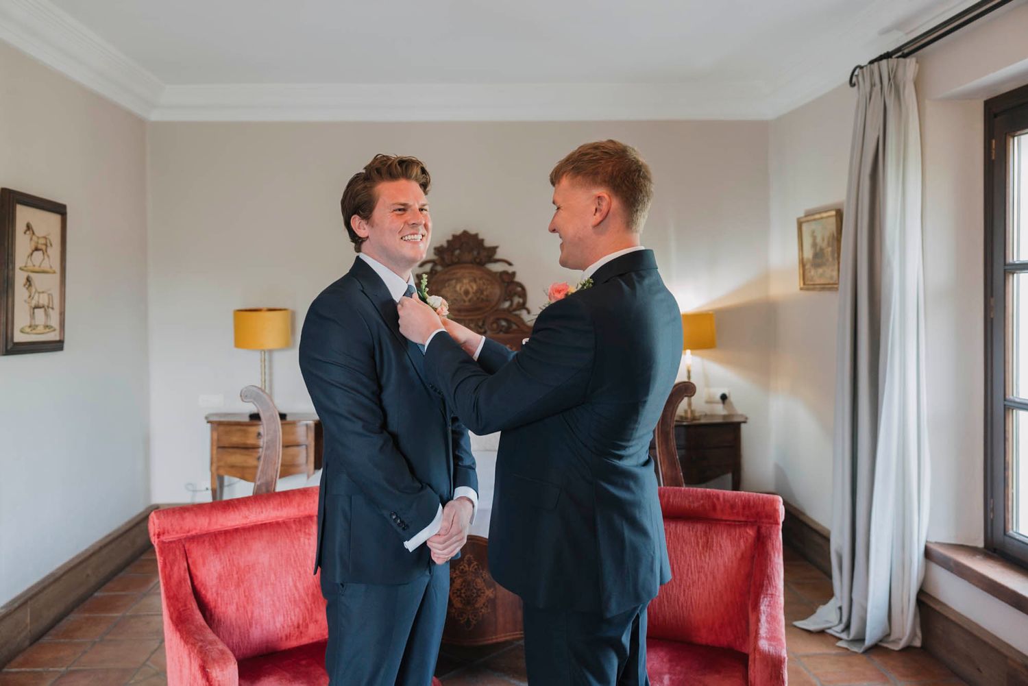 Two people in navy suits helping each other with formal attire adjustments in a room with red furniture.