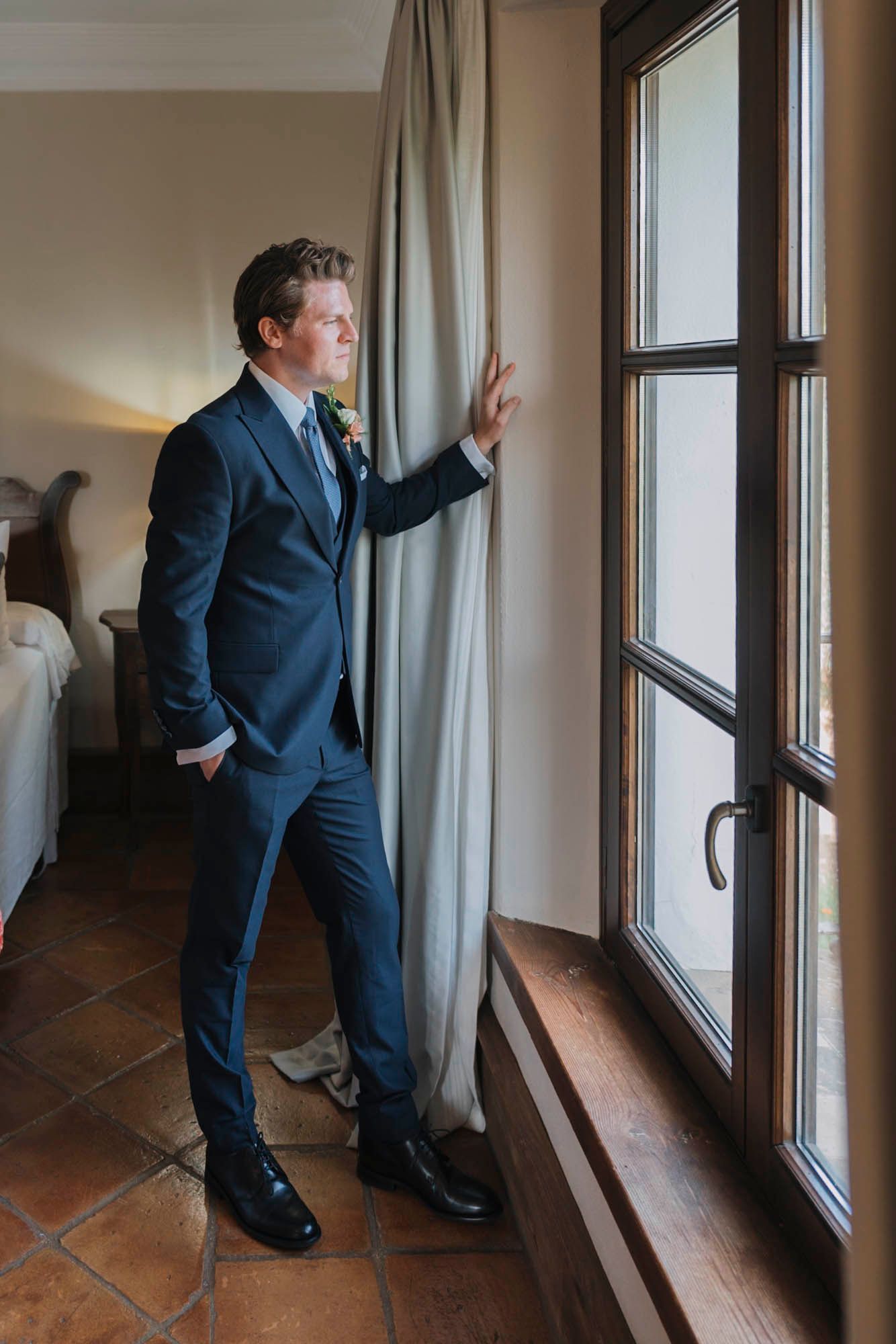 Person in a dark blue suit looking through tall window with curtains and terracotta floor tiles.