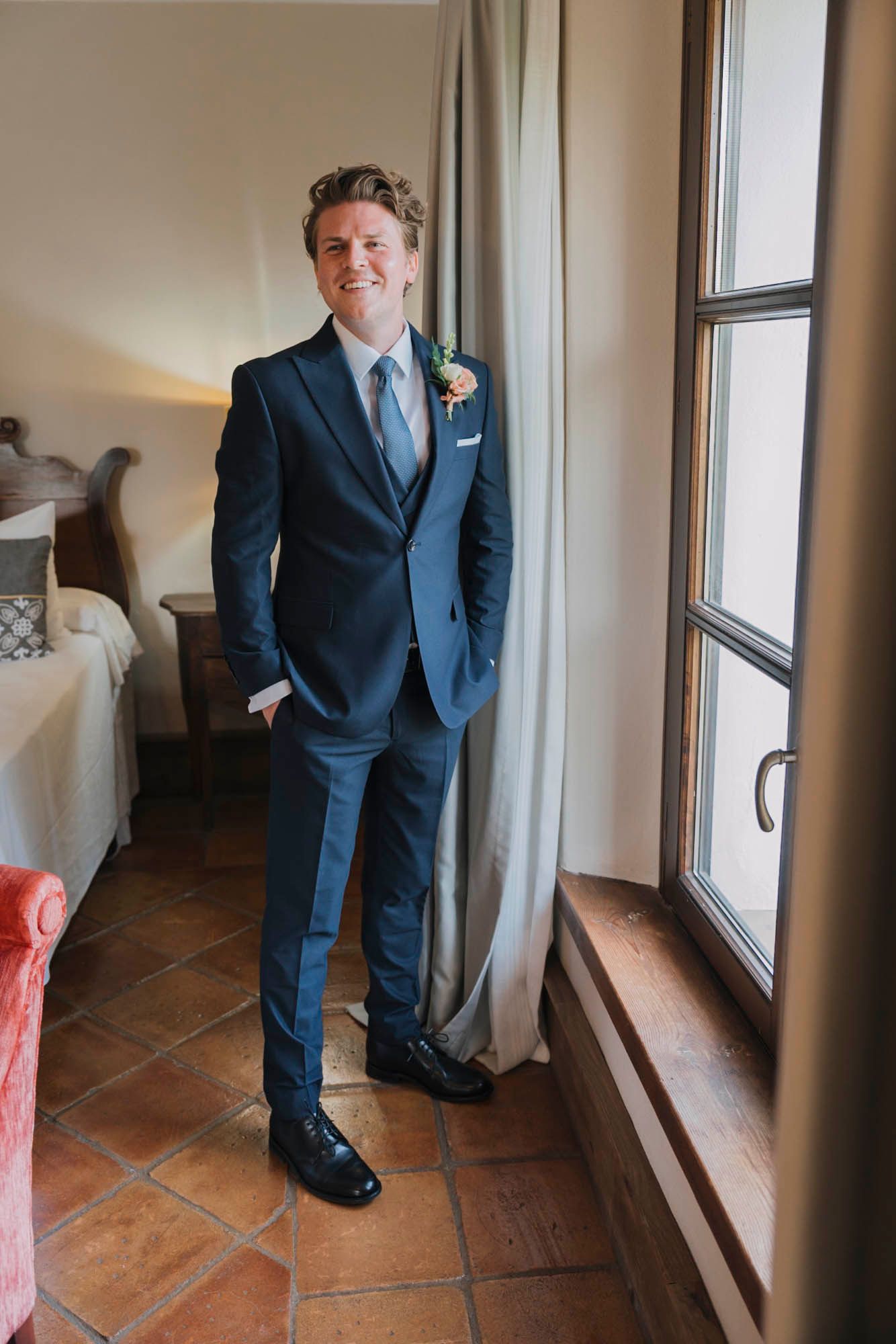 Person in navy suit with boutonniere poses confidently by window in elegant room with terracotta flooring.