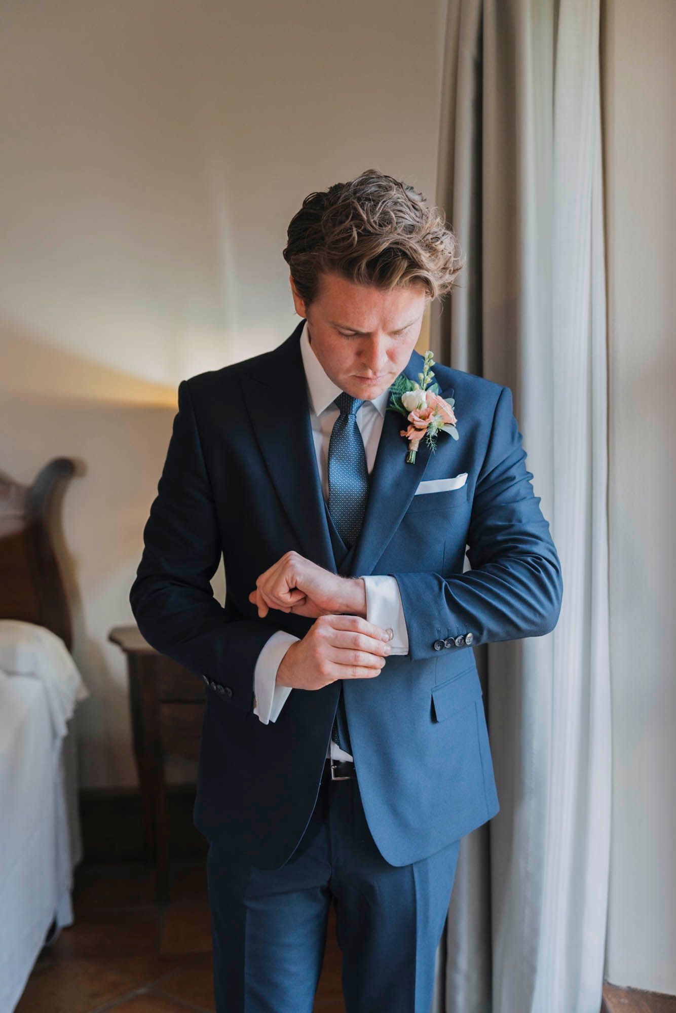 Person in blue suit adjusts sleeve cuff while standing by window in formal wedding attire.