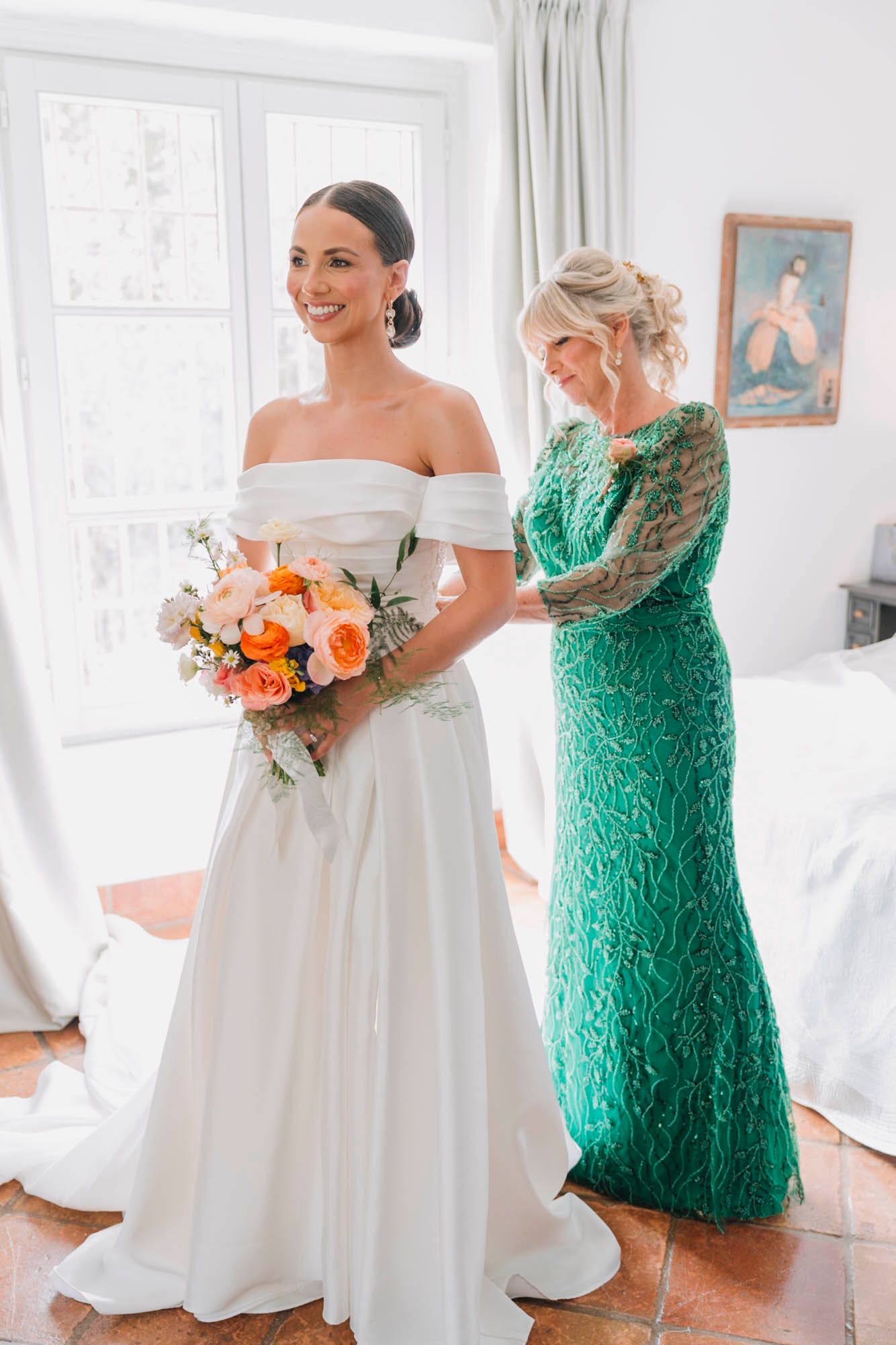 Two people in wedding attire, one in white dress holding coral bouquet and one in emerald green lace gown.