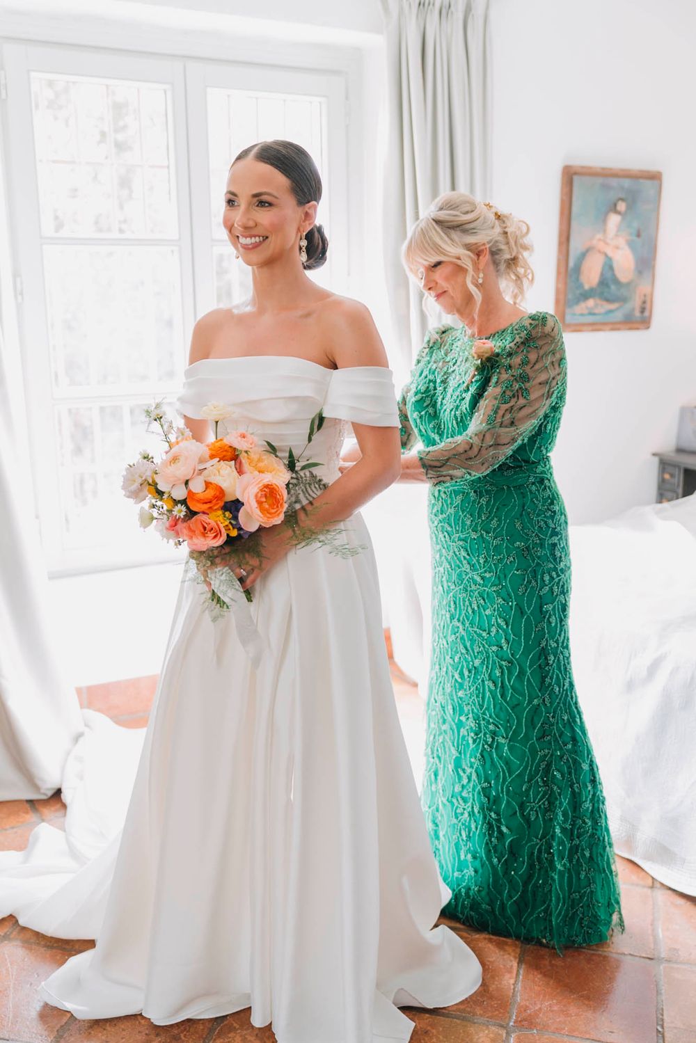 Two people in wedding attire, one in white dress holding coral bouquet and one in emerald green lace gown.