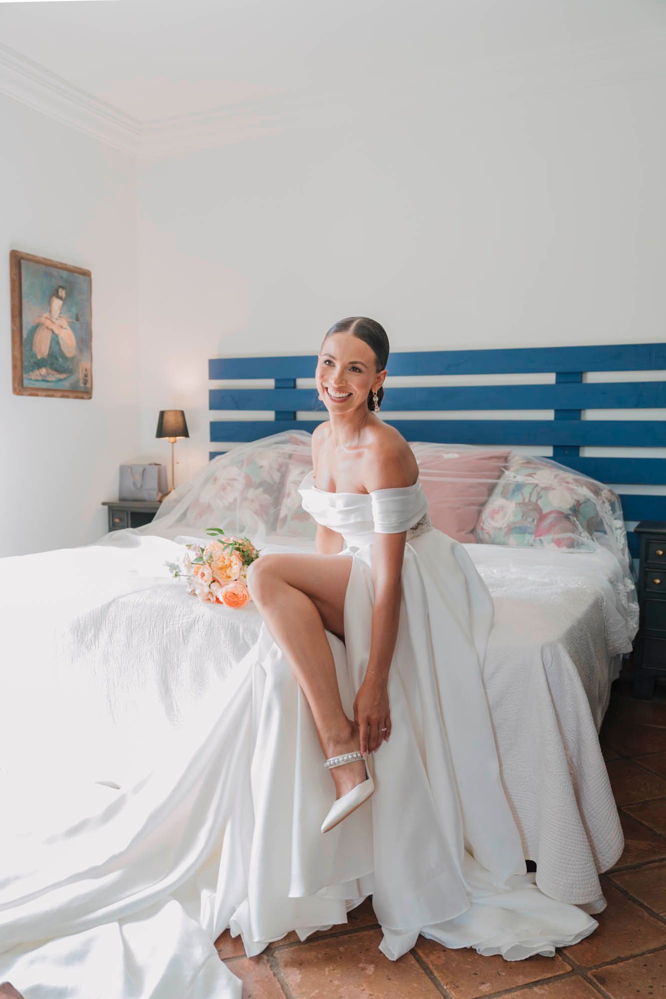 Person in off-shoulder white wedding dress sits elegantly on bed with coral bouquet.