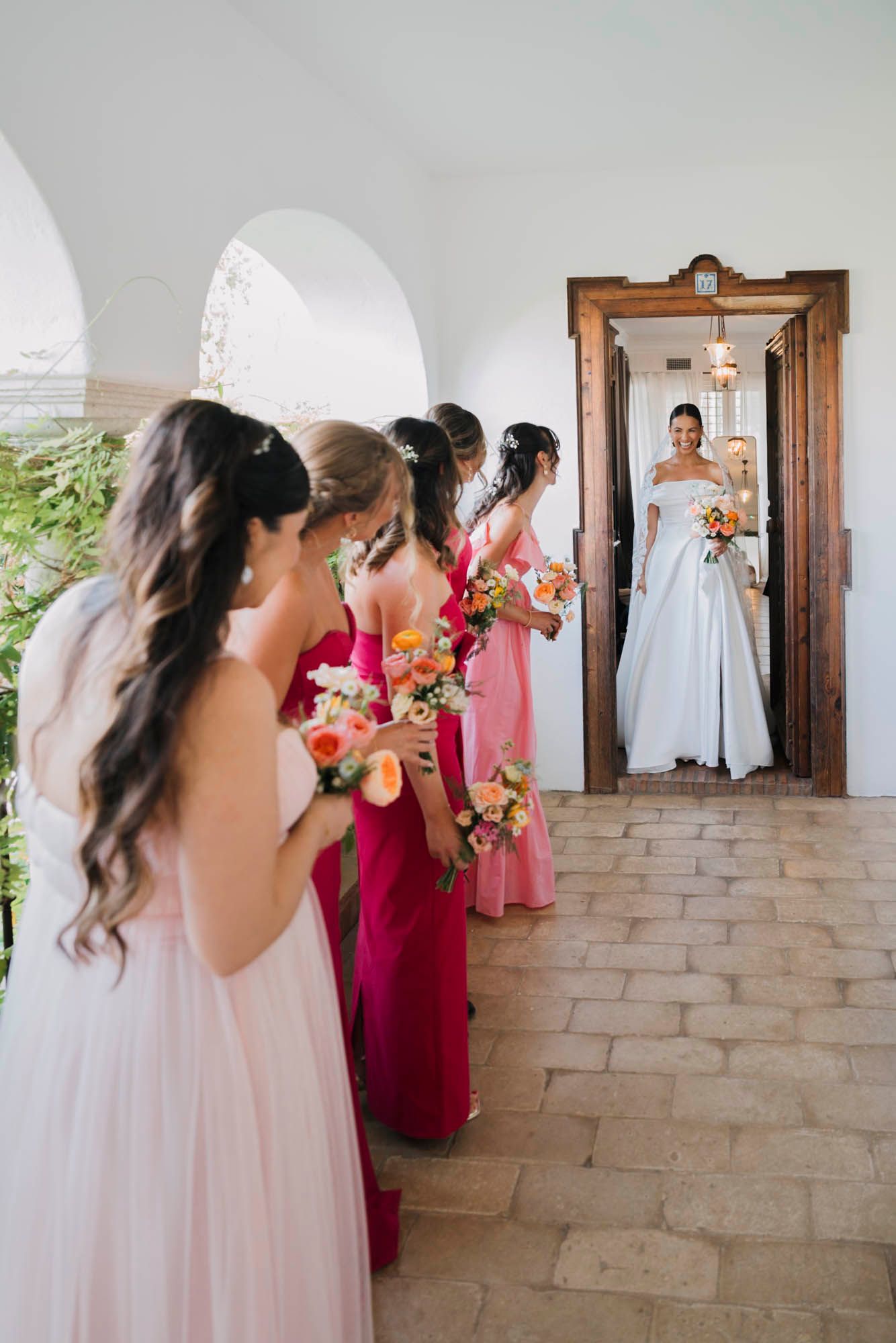 Bridesmaids in pink dresses watch bride in doorway during wedding preparation.