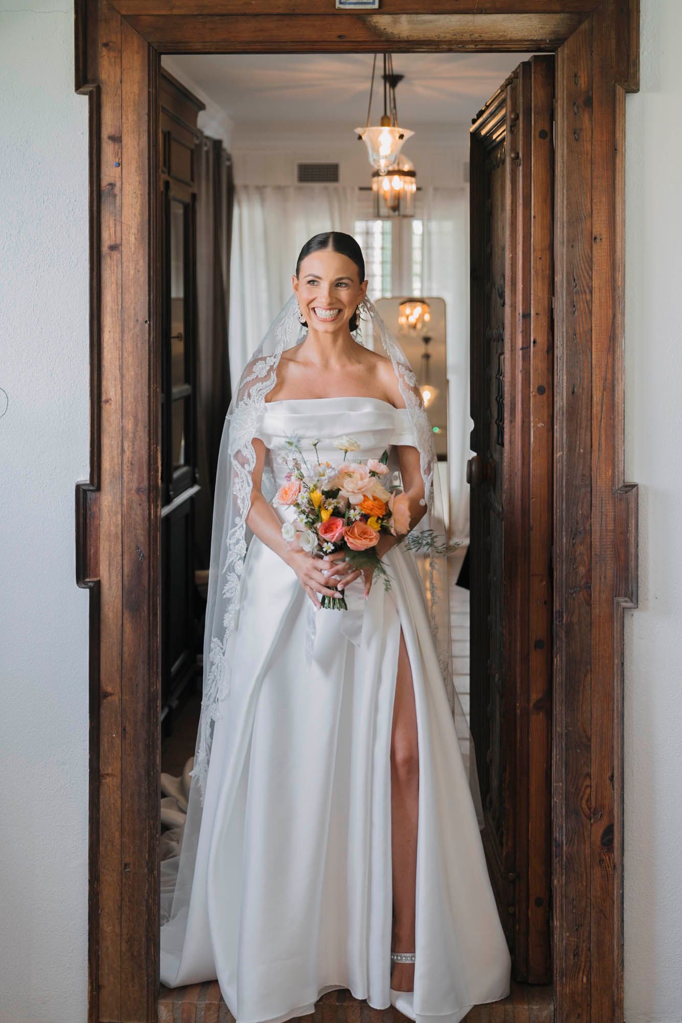 Person in elegant white wedding gown holding colorful bouquet poses in wooden doorframe.