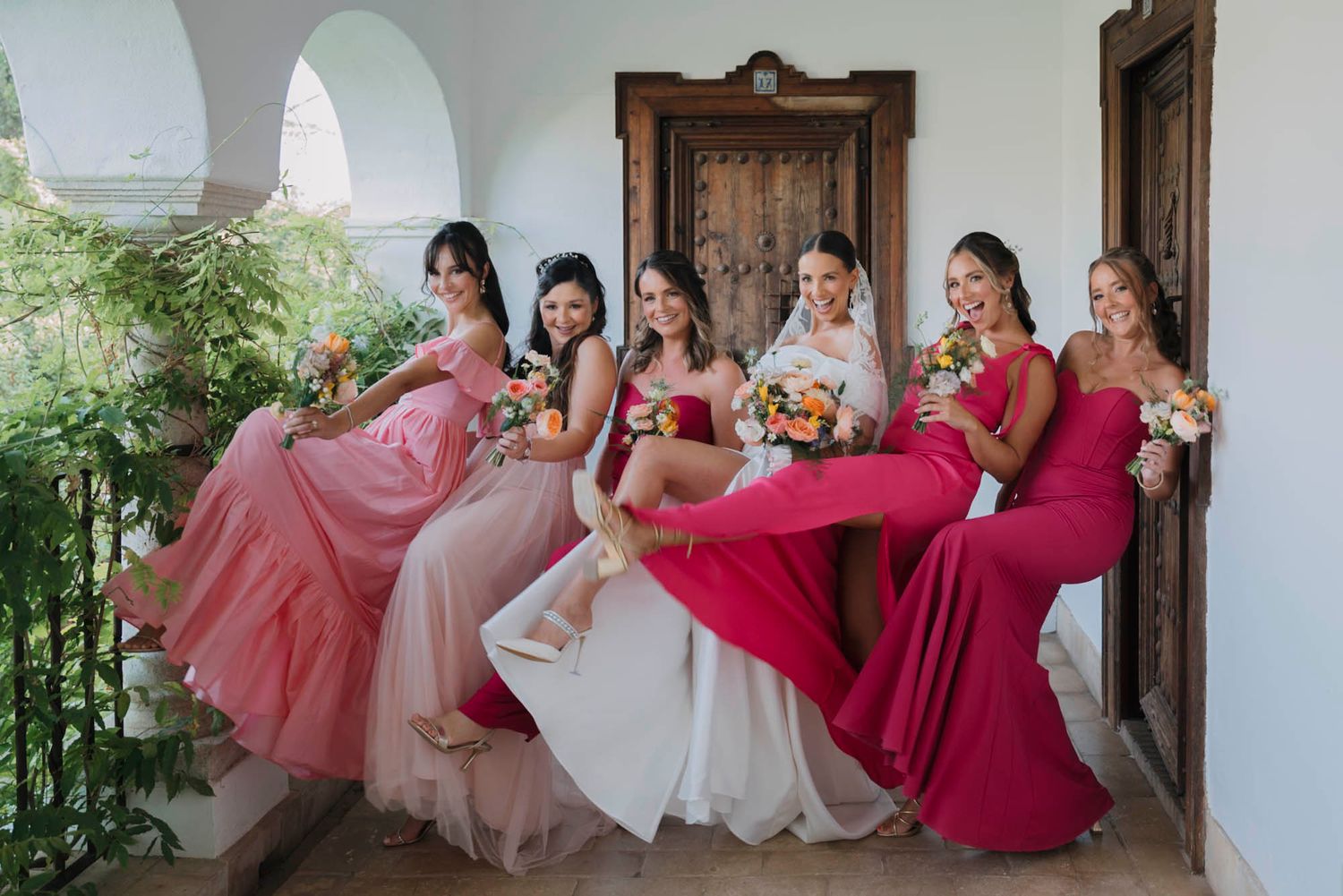 Bridesmaids in various shades of pink and red dresses pose playfully against white archway.
