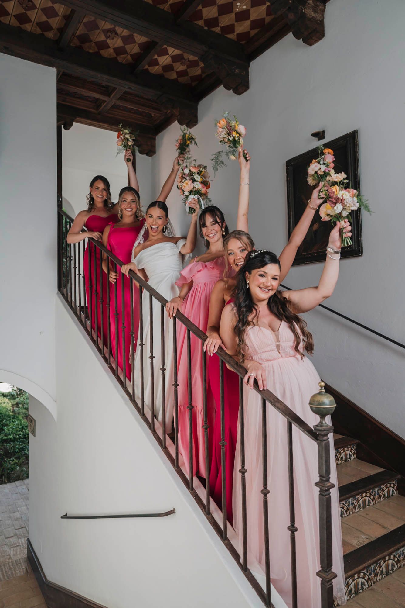Bridesmaids in shades of pink and red dresses pose with bouquets on an elegant staircase.