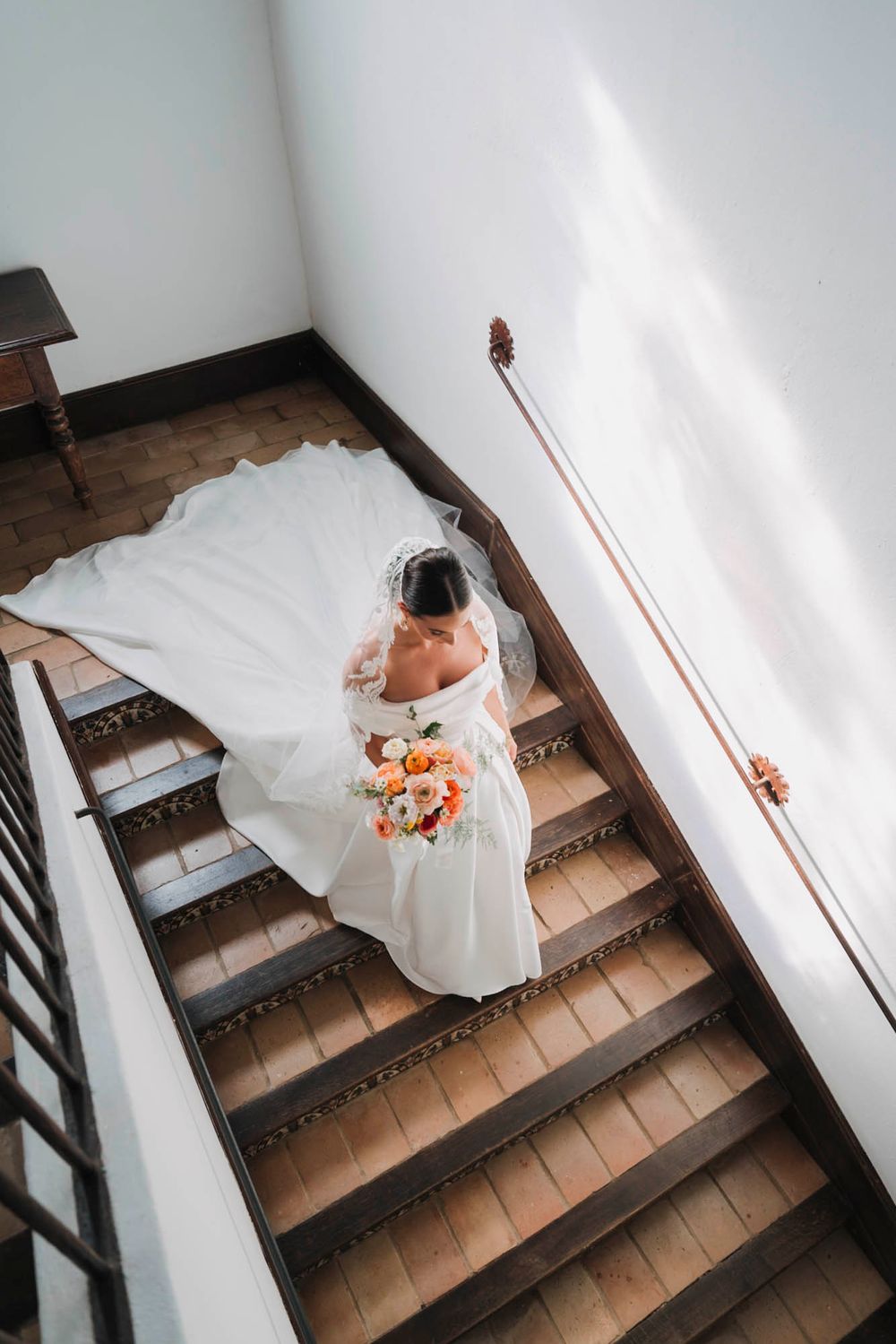 Aerial view of bride in white dress holding bouquet while descending wooden staircase.