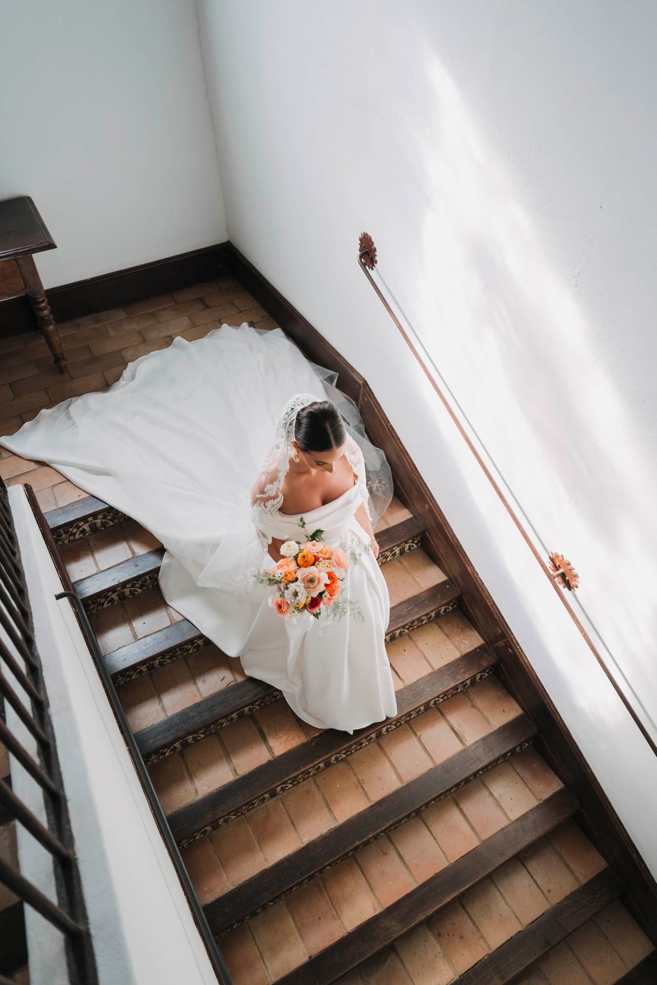 Aerial view of bride in white dress holding bouquet while descending wooden staircase.