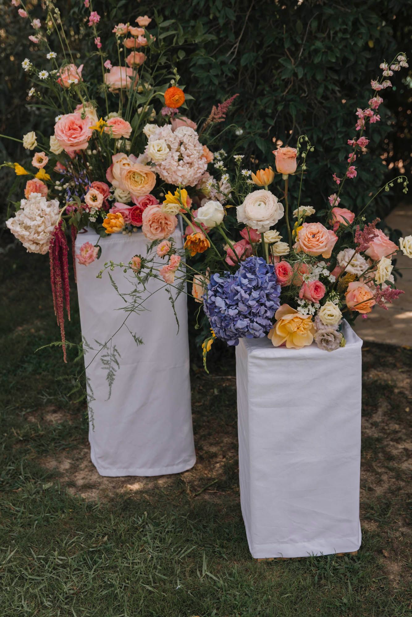 Two white pedestals displaying lush floral arrangements with roses and hydrangeas in garden setting.