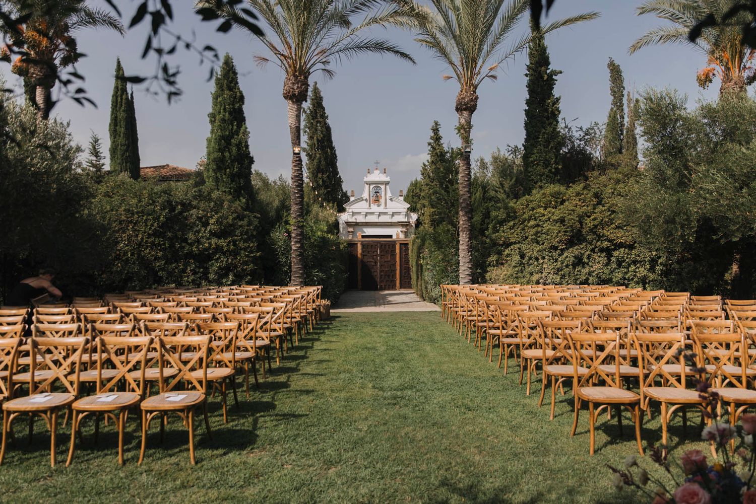 Mediterranean wedding venue with palm trees and wooden chairs leading to white gazebo.