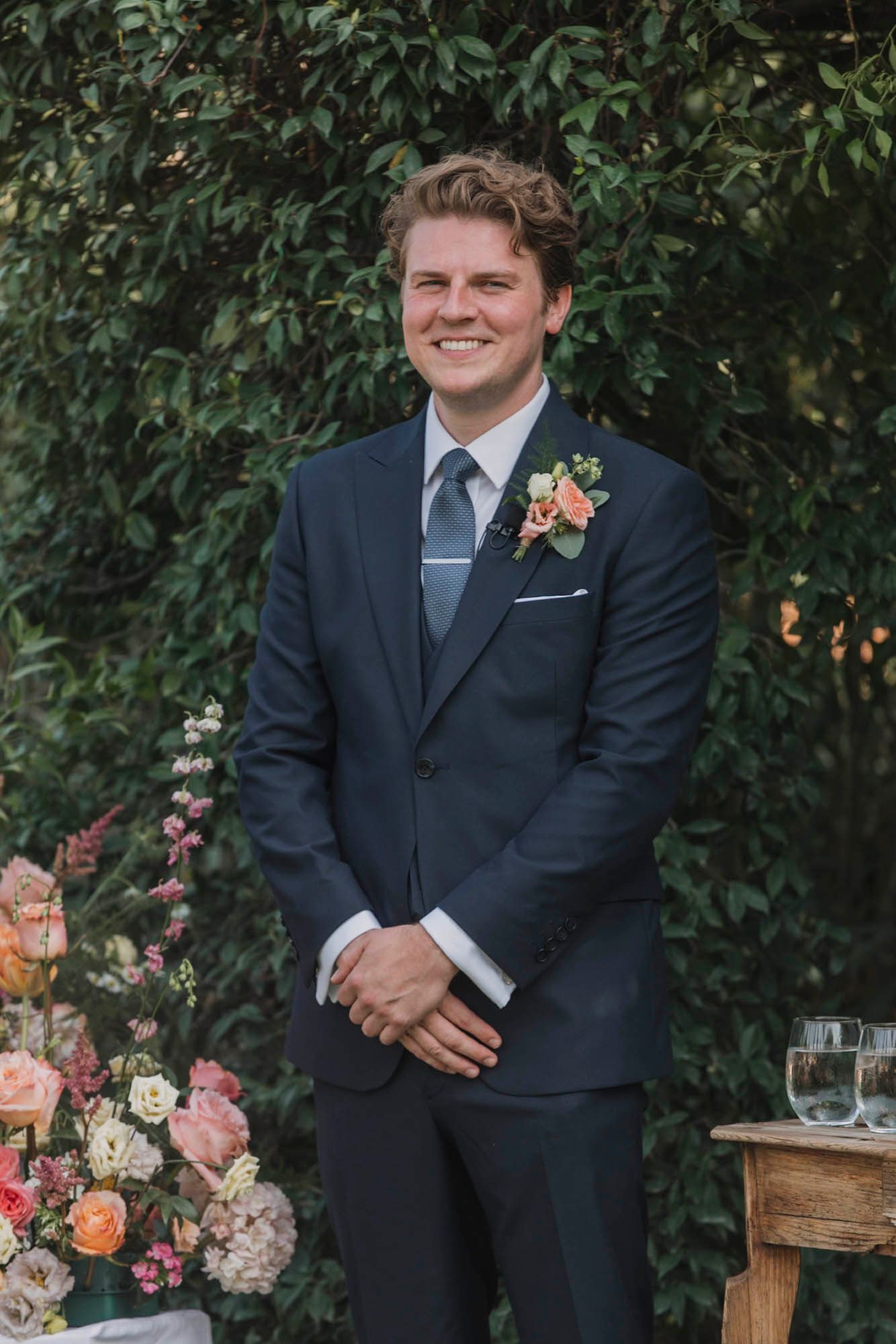 Groom in navy blue suit with boutonniere stands against green foliage backdrop.