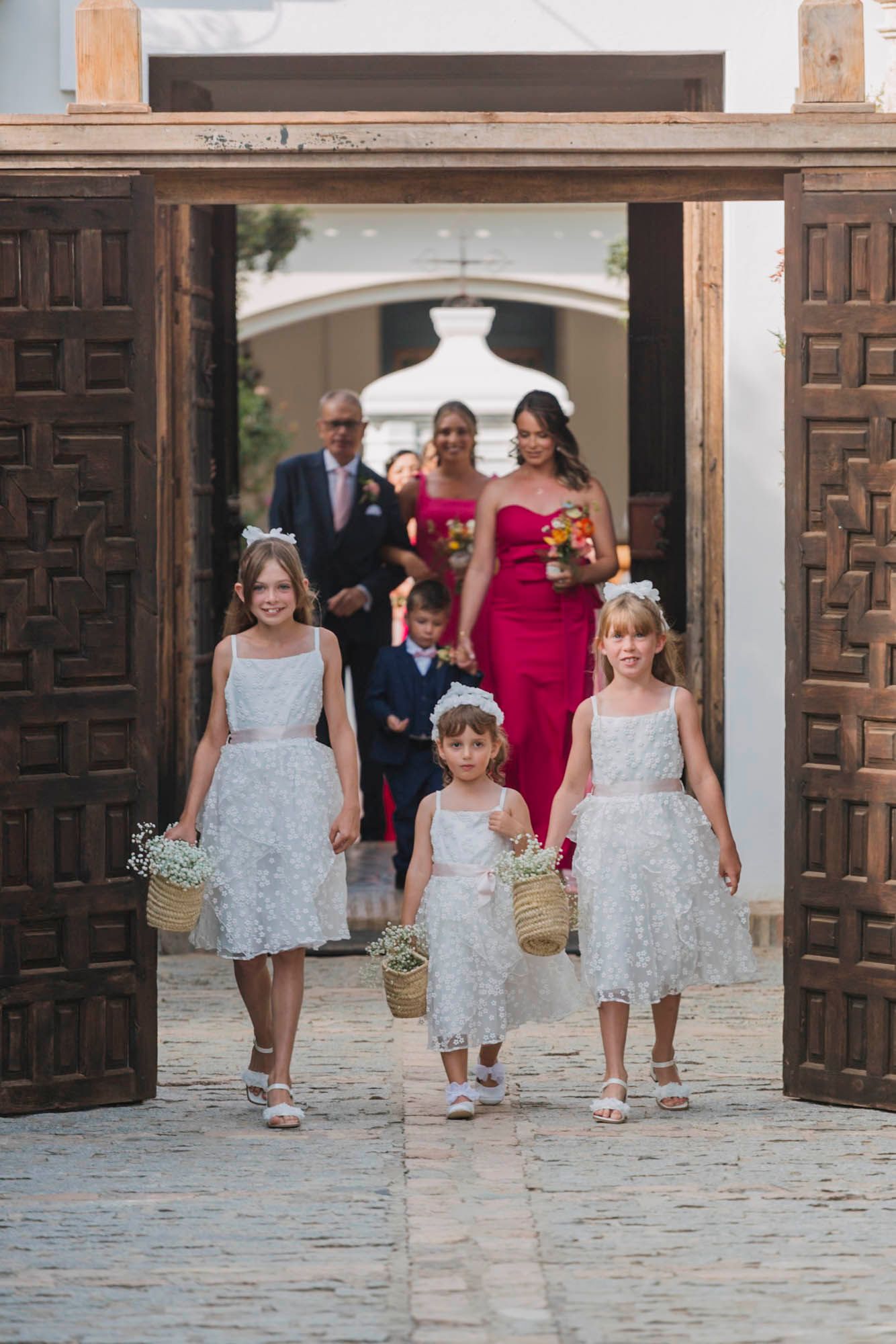 Flower girls in white lacy dresses lead wedding party through ornate wooden doorway.