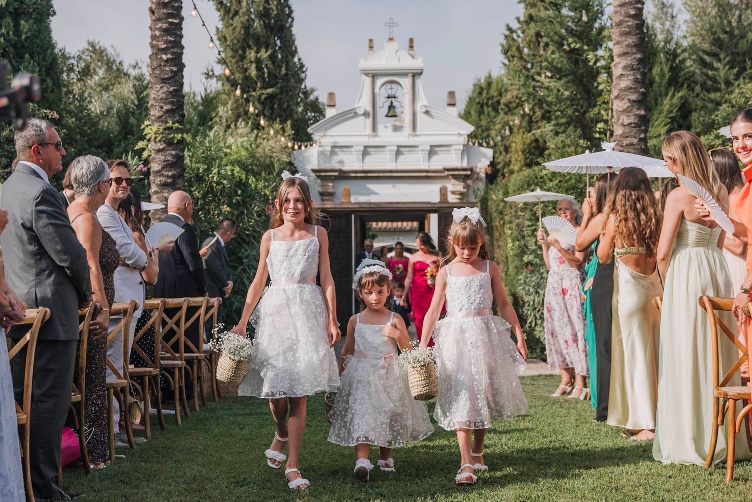 Wedding procession walks down outdoor aisle with guests seated on wooden chairs.