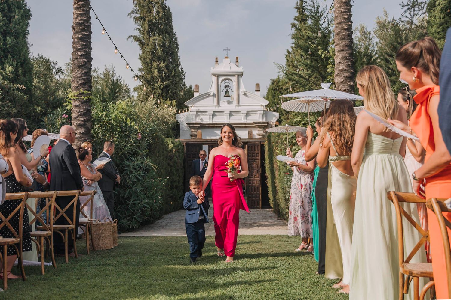 Wedding ceremony processional with guests in colorful attire seated along garden aisle.