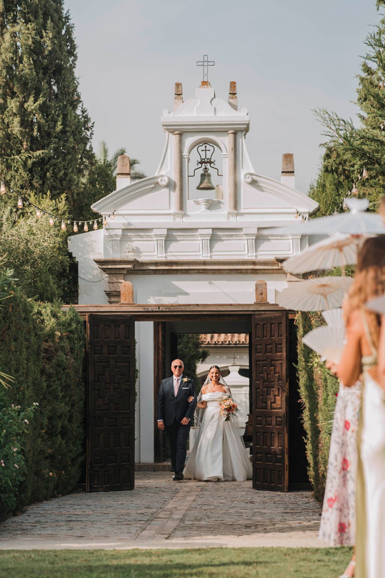 A couple exits through ornate white church gates with a bell tower during their wedding ceremony.