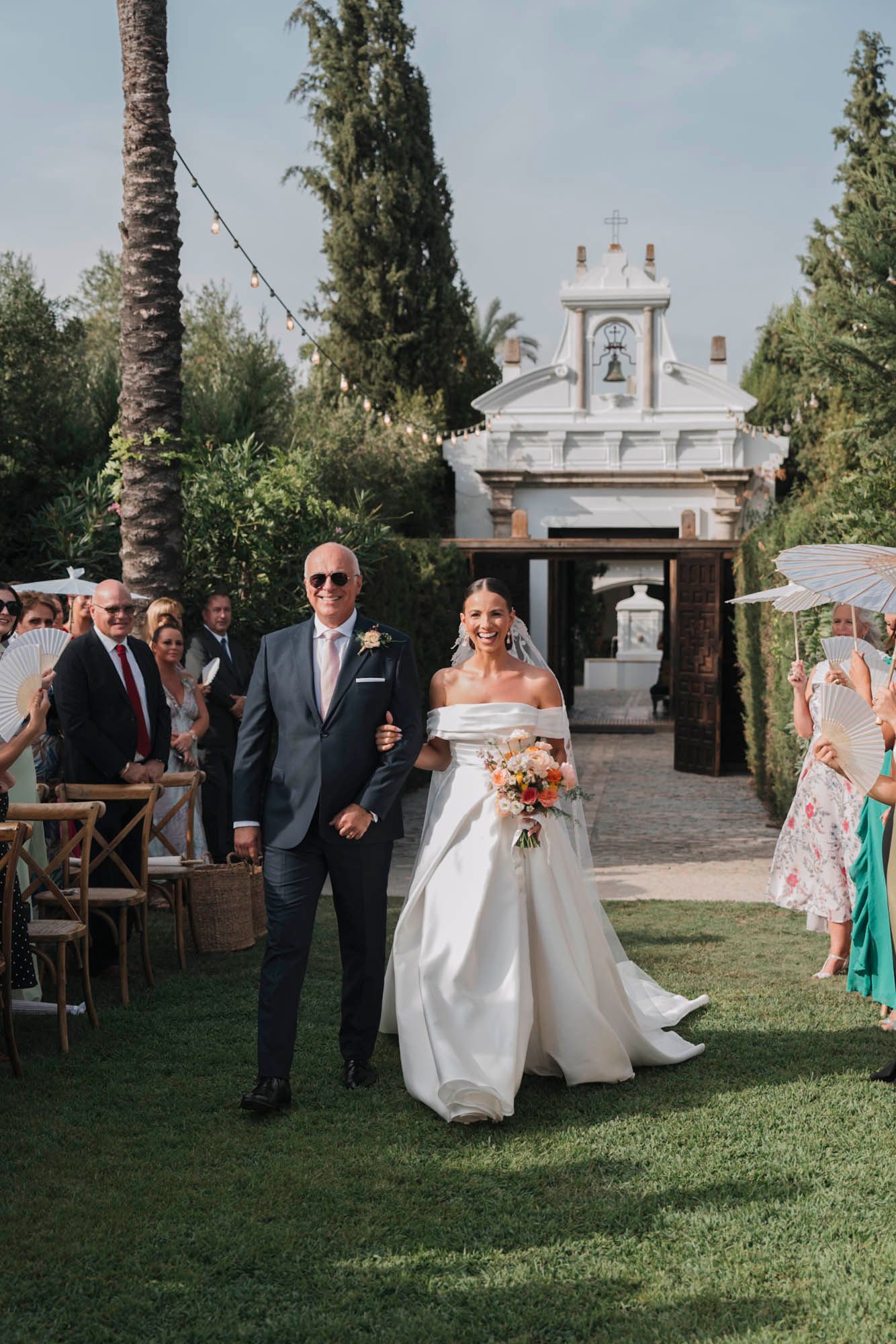 Wedding party walks down an outdoor aisle with cypress trees and palm trees in a garden setting.