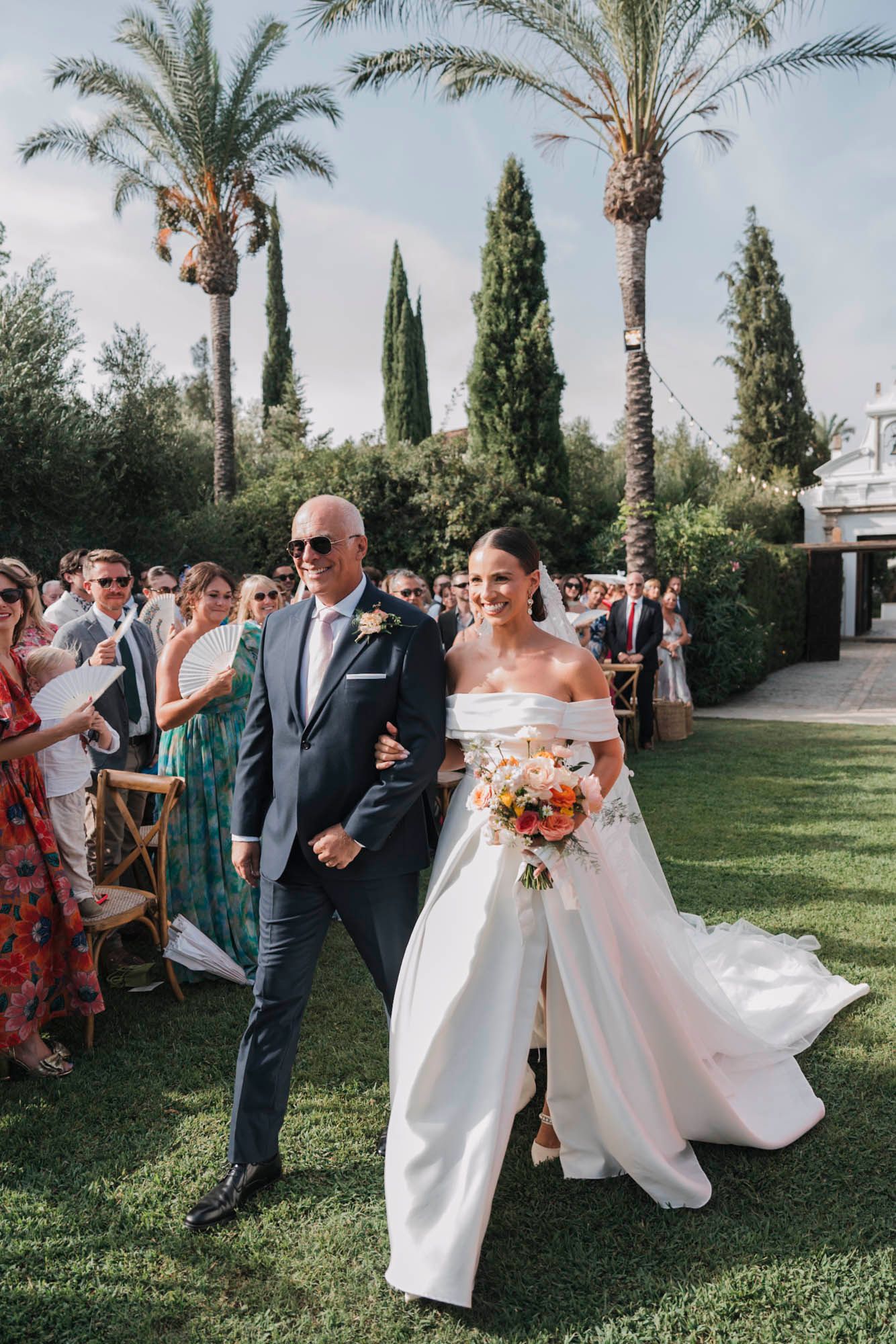 Wedding processional on a manicured lawn lined with palm trees and cypress trees.