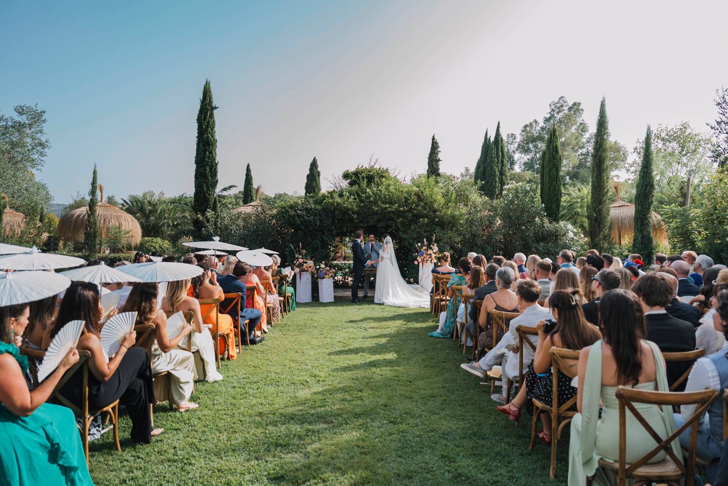 Outdoor wedding ceremony with guests seated on white chairs on a manicured lawn.