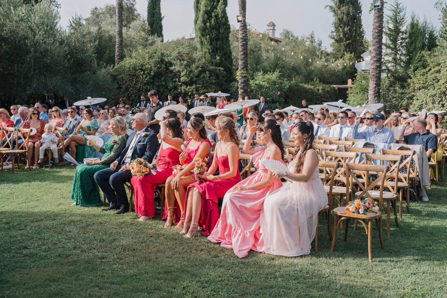 Wedding guests in colorful formal attire seated on wooden chairs during an outdoor garden ceremony.