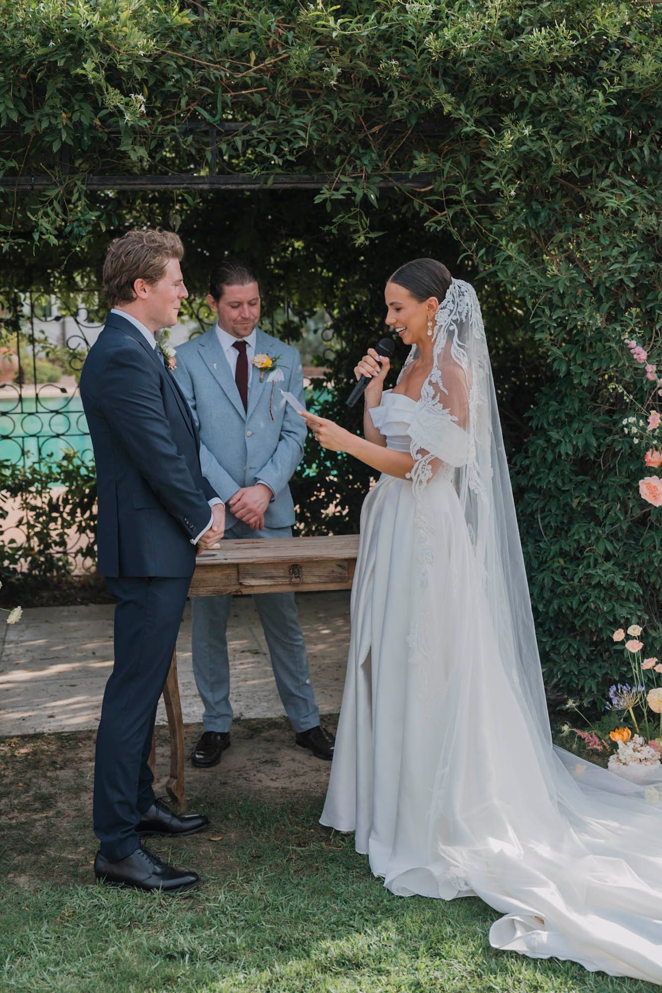 Wedding ceremony taking place under a lush garden archway with formal attire and traditional decor.