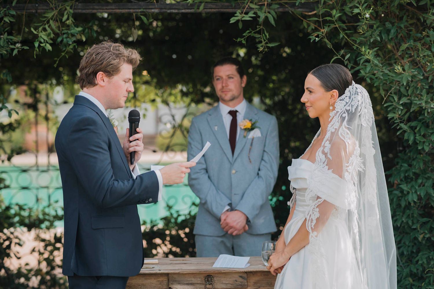 Intimate outdoor wedding ceremony moment with participants exchanging vows under a green archway.